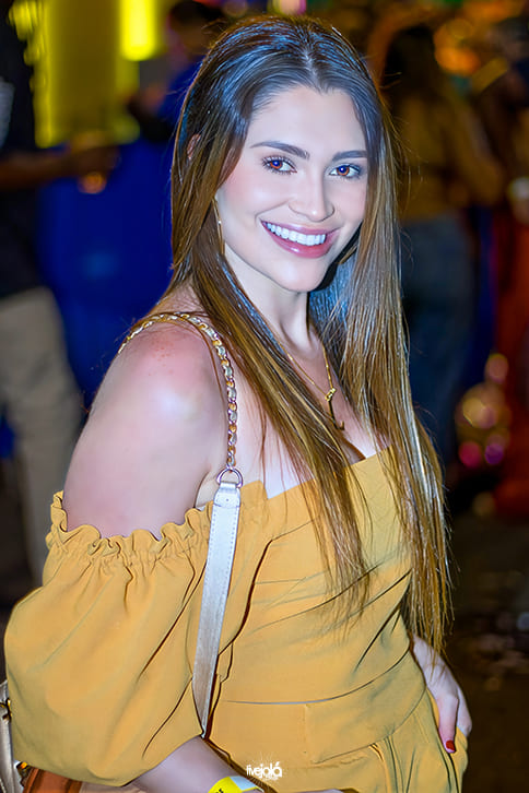 The Music Beach - Me Leva Festival - A imagem mostra uma mulher sorrindo Ela está usando um vestido amarelo mostarda, com ombros de fora e mangas bufantes, além de uma alça de bolsa dourada com detalhe em corrente. Seus cabelos são lisos, longos e castanho-claros, caindo sobre um dos ombros. 