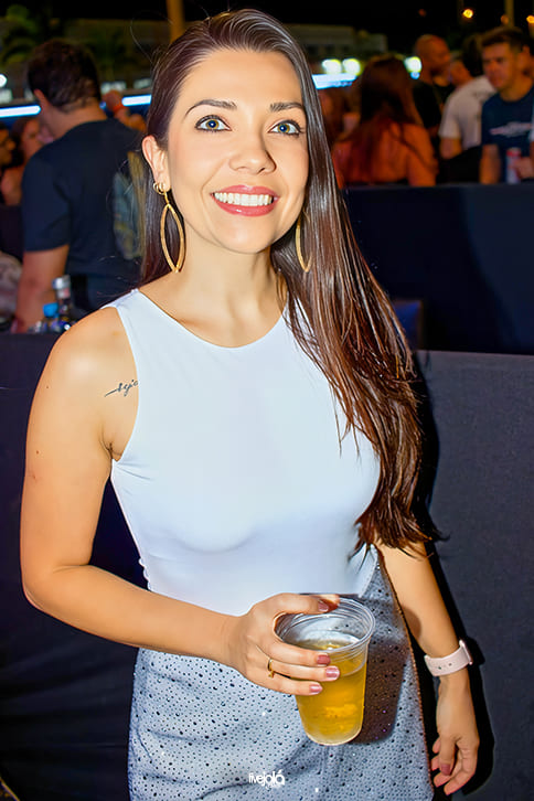 The Music Beach - Me Leva Festival - A imagem mostra uma mulher sorrindo  Ela tem cabelos longos e lisos, castanhos, e usa brincos grandes dourados em formato de argola. Está vestindo uma blusa branca sem mangas e uma saia ou shorts prateado com brilhos. No braço esquerdo, ela usa um relógio ou smartwatch claro, e segura um copo plástico com bebida dourada, provavelmente cerveja.