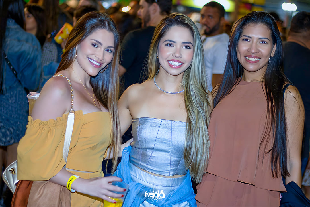 The Music Beach - Me Leva Festival - A imagem mostra três mulheres sorrindo Da esquerda para a direita:  Mulher com vestido amarelo mostarda, ombros de fora e alças com detalhes dourados; segura um copo amarelo transparente.  Mulher com top prateado tomara que caia, colar prateado e uma camisa azul amarrada na cintura.  Mulher com blusa marrom de alças finas e cabelos longos lisos.
