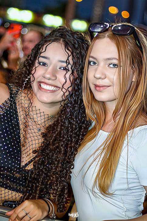 	 The Music Beach - Me Leva Festival - A imagem mostra duas jovens sorrindo e posando juntas para a foto A jovem à esquerda tem cabelo cacheado e volumoso, usa um vestido preto com detalhes brilhantes e uma rede transparente no decote, transmitindo um visual festivo. Ela sorri amplamente, mostrando os dentes, e segura a grade à sua frente.  A jovem à direita tem cabelos longos e lisos, de cor ruiva, e veste uma blusa branca. Ela mantém um sorriso mais sutil e usa óculos de sol apoiados sobre a cabeça.