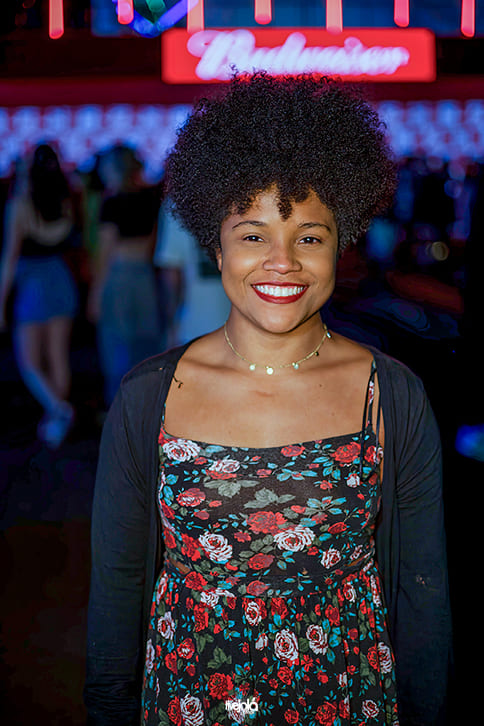Festival Timbre - A imagem mostra uma mulher. Ela tem cabelo crespo volumoso em estilo black power e está usando um vestido estampado com flores vermelhas e verdes sobre fundo preto. Por cima, veste um cardigã preto leve. No pescoço, usa um colar delicado com pequenos pingentes dourados.  A iluminação destaca seu rosto e o vestido.