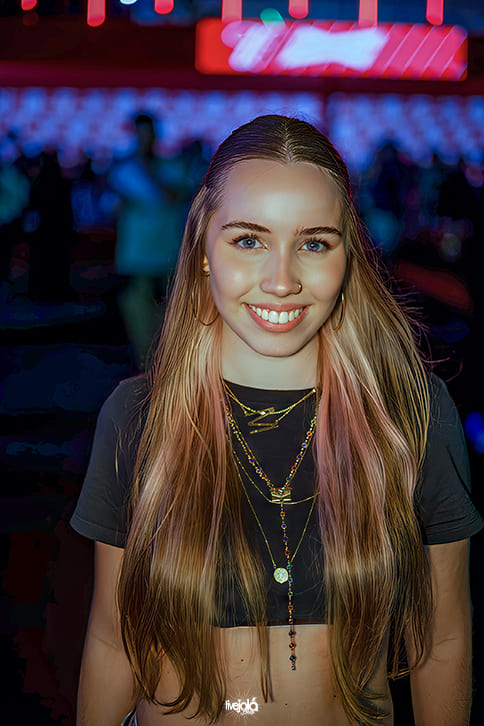 Festival Timbre - A imagem mostra uma mulher sorridente. Ela tem cabelos loiros longos e lisos, com leves mechas em tons mais claros e rosados. Usa uma blusa preta curta, deixando parte da barriga à mostra, e acessórios dourados: colares em camadas, incluindo um com pingente em forma de pássaro e outro com detalhes coloridos. Ela também usa argolas douradas e um piercing discreto no nariz.