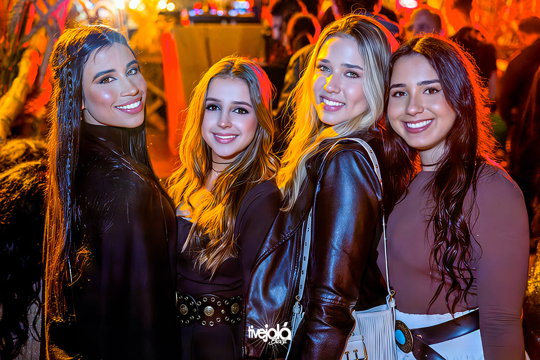 Quatro mulheres, um mulher de cabelos pretos encaracolados sorrindo, uma mulher loira de cabelos longos sorrindo, uma mulher loira cabelos longos sorrindo e outra cabelos encaracolados castanhos escuros