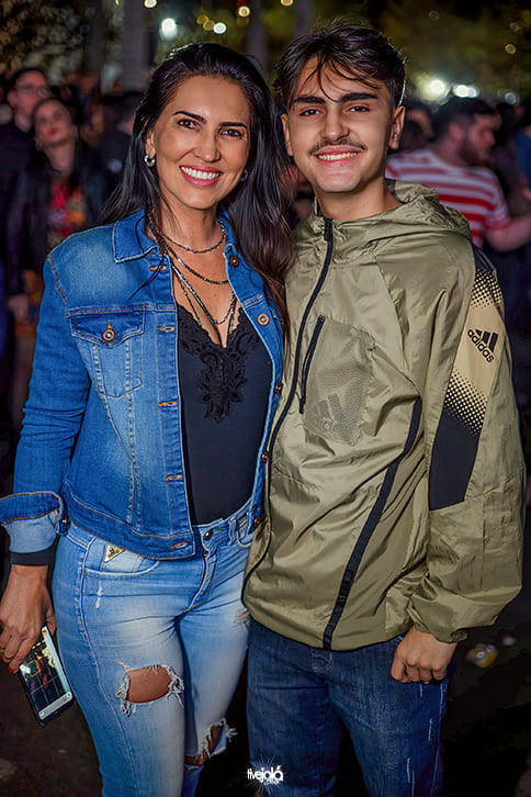 Festival Timbre - A imagem mostra uma mulher e um jovem posando juntos. A mulher, à esquerda, está sorridente, usando uma jaqueta jeans azul sobre uma blusa preta rendada e calça jeans clara com rasgo no joelho. Ela segura um celular na mão direita e usa colares pretos.  O jovem, à direita, também sorri para a câmera, vestindo uma jaqueta esportiva verde da Adidas com detalhes pretos e calça jeans escura.