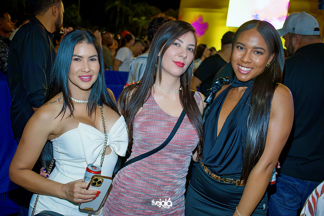 The Music Beach - Me Leva Festival - A imagem mostra três mulheres sorrindo e posando para a câmera em um evento noturno.  A mulher à esquerda tem cabelo liso e escuro, veste um vestido branco tomara que caia com um grande laço na parte frontal, segura uma lata de bebida e um celular com capa dourada. Ela usa colar de pérolas e bolsa de corrente dourada.  A mulher ao centro tem cabelo liso e castanho-escuro, veste um vestido justo em tons de cinza e rosa, usa brincos grandes de argola e carrega uma bolsa transversal preta.  A mulher à direita tem cabelo liso e comprido, veste um vestido preto com decote profundo e detalhe de laço no ombro, com cinto dourado.  Ao fundo, há várias pessoas e iluminação colorida, sugerindo que se trata de uma festa ou show.