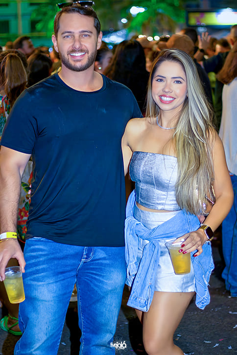 The Music Beach - Me Leva Festival - A imagem mostra um homem e uma mulher posando juntos O homem, à esquerda, veste uma camiseta preta e calça jeans azul, usando óculos escuros apoiados na cabeça. Ele segura um copo com bebida amarela na mão direita e sorri para a câmera.  A mulher, à direita, veste um conjunto prateado composto por top tomara-que-caia e shorts curtos. Ela tem uma blusa azul clara amarrada na cintura, usa relógio preto no pulso esquerdo e segura um copo semelhante ao do homem na mão direita. Seus cabelos são longos e loiros, e ela sorri para a câmera.