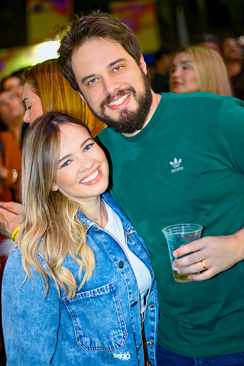 The Music Beach - Me Leva Festival -  A imagem mostra um casal sorrindo A mulher, à esquerda, tem cabelo loiro com raiz mais escura, veste camiseta branca e jaqueta jeans azul clara. Ela sorri amplamente, inclinando levemente a cabeça para o homem.  O homem, à direita, tem cabelo castanho e barba cheia, veste camiseta verde-escura da marca Adidas e segura um copo plástico transparente com bebida. Ele abraça a mulher pelos ombros.