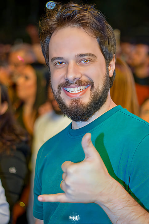 The Music Beach - Me Leva Festival - A imagem mostra um homem sorridente Ele tem cabelo castanho e barba cheia, veste uma camiseta verde-escura e olha diretamente para a câmera.  Com a mão direita, faz o gesto “hang loose” (polegar e mindinho estendidos, os outros dedos dobrados).
