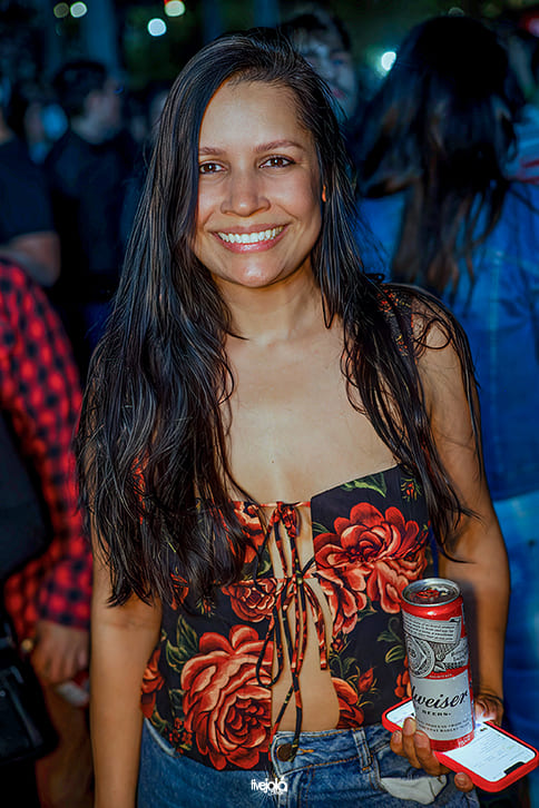 Festival Timbre - A imagem mostra uma mulher sorridente. Ela tem cabelos longos, lisos e escuros, soltos sobre os ombros. Veste um top estampado com grandes flores vermelhas e folhagens verdes, amarrado na parte frontal por cordões, e uma calça jeans azul.  Na mão direita, segura uma lata de cerveja Budweiser e usando o celular como suporte para bebida.