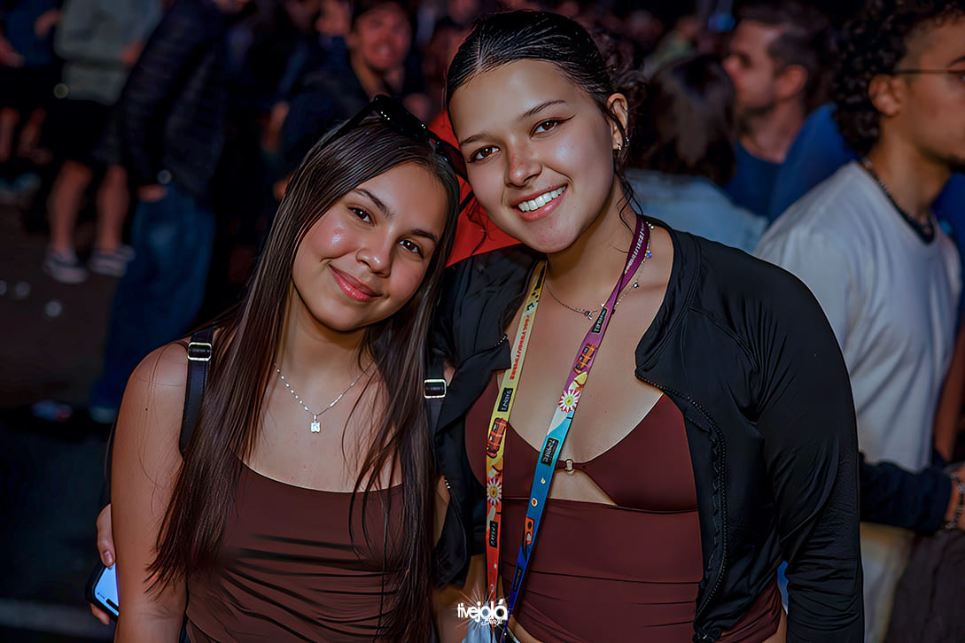 Festival Timbre - A imagem mostra duas mulheres jovens posando juntas e sorrindo. A mulher à esquerda tem cabelos lisos e longos, veste um top marrom sem mangas e um colar delicado com pingente quadrado. Ela segura um celular na mão direita. A mulher à direita também veste um top marrom, combinado com um casaco preto leve aberto, e usa um cordão colorido com crachá pendurado no pescoço.