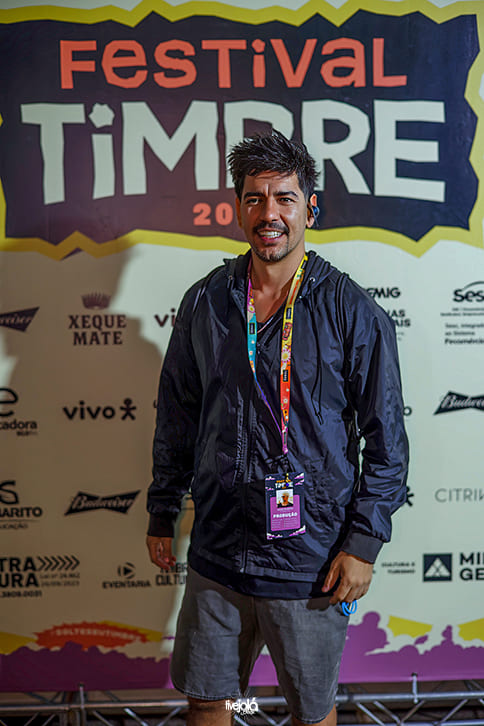 Festival Timbre - A imagem mostra um homem posando sozinho em frente ao painel de patrocinadores do Festival Timbre 2025.  Ele veste uma jaqueta preta com zíper, bermuda cinza e crachá do evento pendurado no pescoço por uma fita colorida. Está sorrindo e olhando diretamente para a câmera.  O fundo é claro, com o logotipo grande e colorido do festival na parte superior e diversos logos de patrocinadores espalhados ao redor.