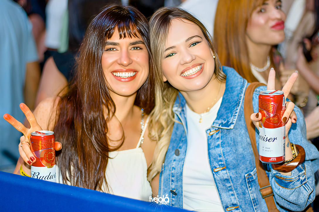 The Music Beach - Me Leva Festival -  A imagem mostra duas mulheres sorrindo amplamente e posando para a câmera Ambas estão segurando latas de cerveja Budweiser e fazendo o gesto de “paz e amor” com as mãos.  A mulher à esquerda tem cabelo castanho longo com franja, usa um vestido ou blusa branca e um colar delicado.  A mulher à direita tem cabelo loiro-claro, veste camiseta branca com jaqueta jeans e carrega uma bolsa marrom de alça lateral.